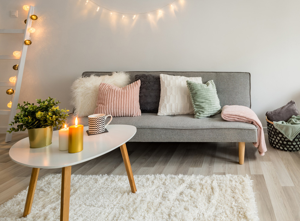 Canapé gris avec coussins aux couleurs pastels, vert, rose, blanc. Une petite table pieds en bois, plateau blanc, un tapis moelleux blanc. Sur la table, une plate, un mug, des bougies allumées. Au mur, une guirlande lumineuse.