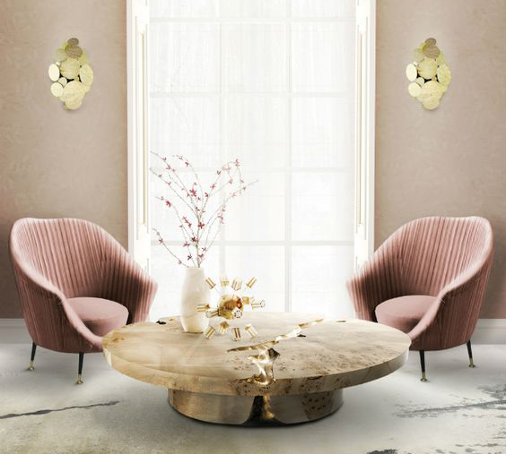 Deux fauteuils identiques en velours vieux rose, encadrant une grande fenêtre. Au centre, une table basse ronde en bois. De chaque côté de la fenêtre, une applique composée de disques dorés est allumée.